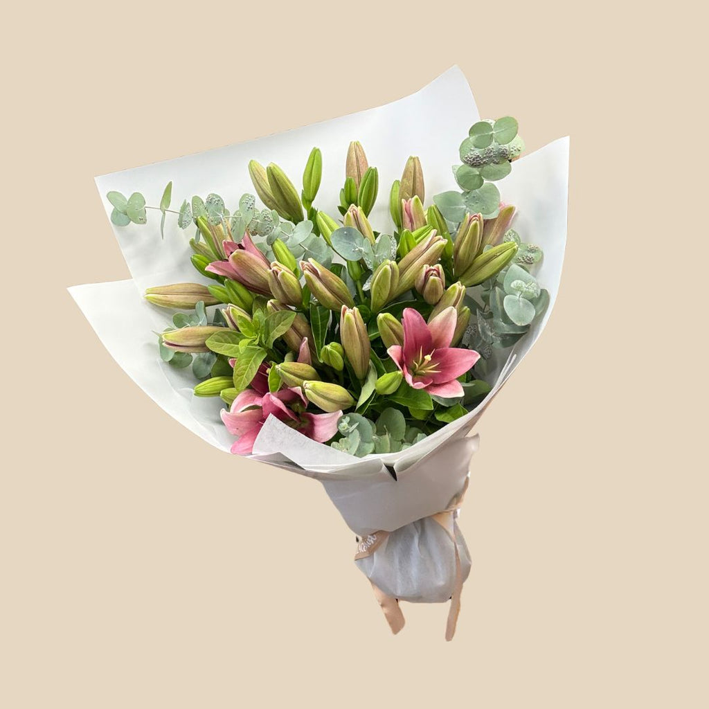 A bouquet of Asiatic Lilies with green leaves, wrapped in white paper and tied with a ribbon.