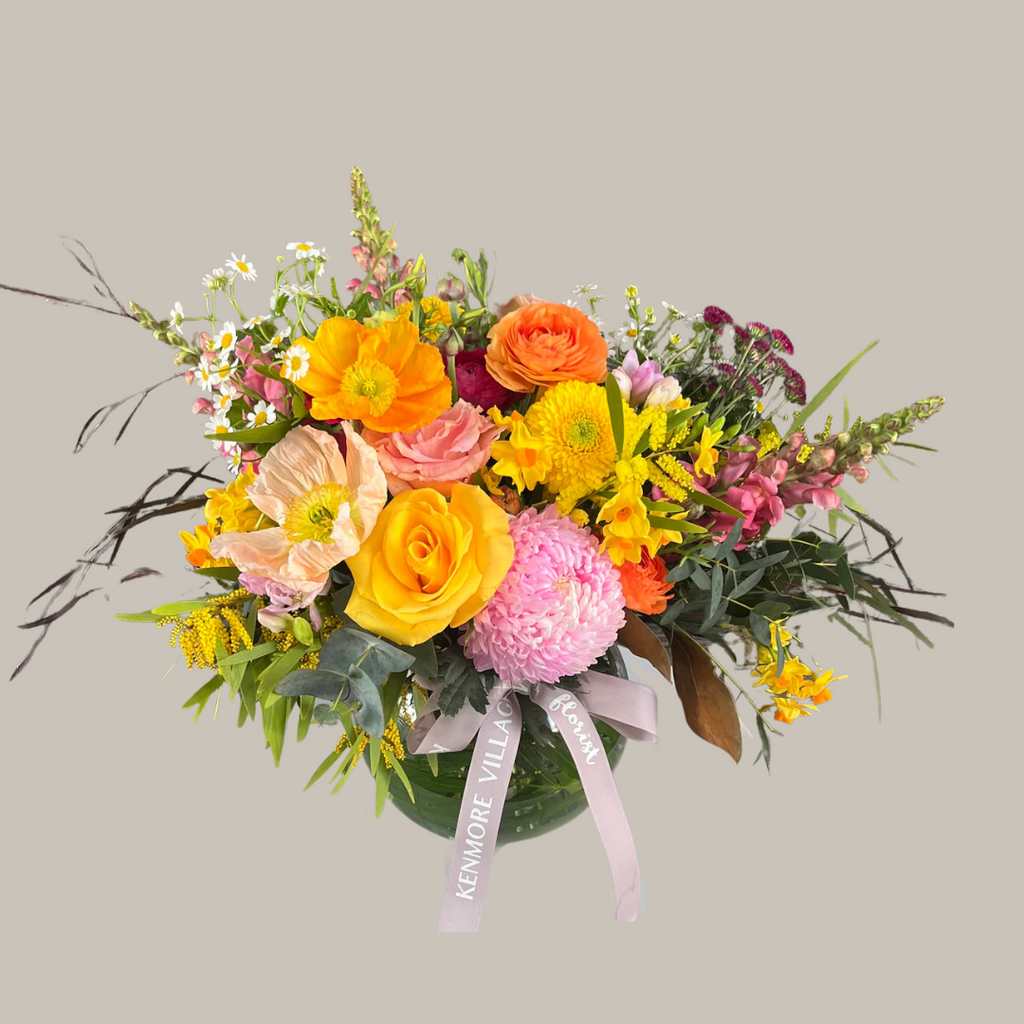 A fishbowl vase arrangement of bright and colorful flowers including yellow, orange, pink, and other colored blooms, with a ribbon decoration. Kenmore Village Florist. Brisbane Same Day Flower Delivery.