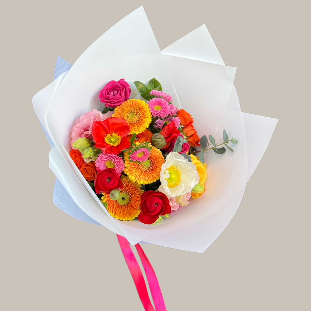 A colorful bouquet of flowers including shades of red, orange, yellow, and green, wrapped in white paper with a pink ribbon. Kenmore Village Florist. Brisbane Florist. Same day delivery.
