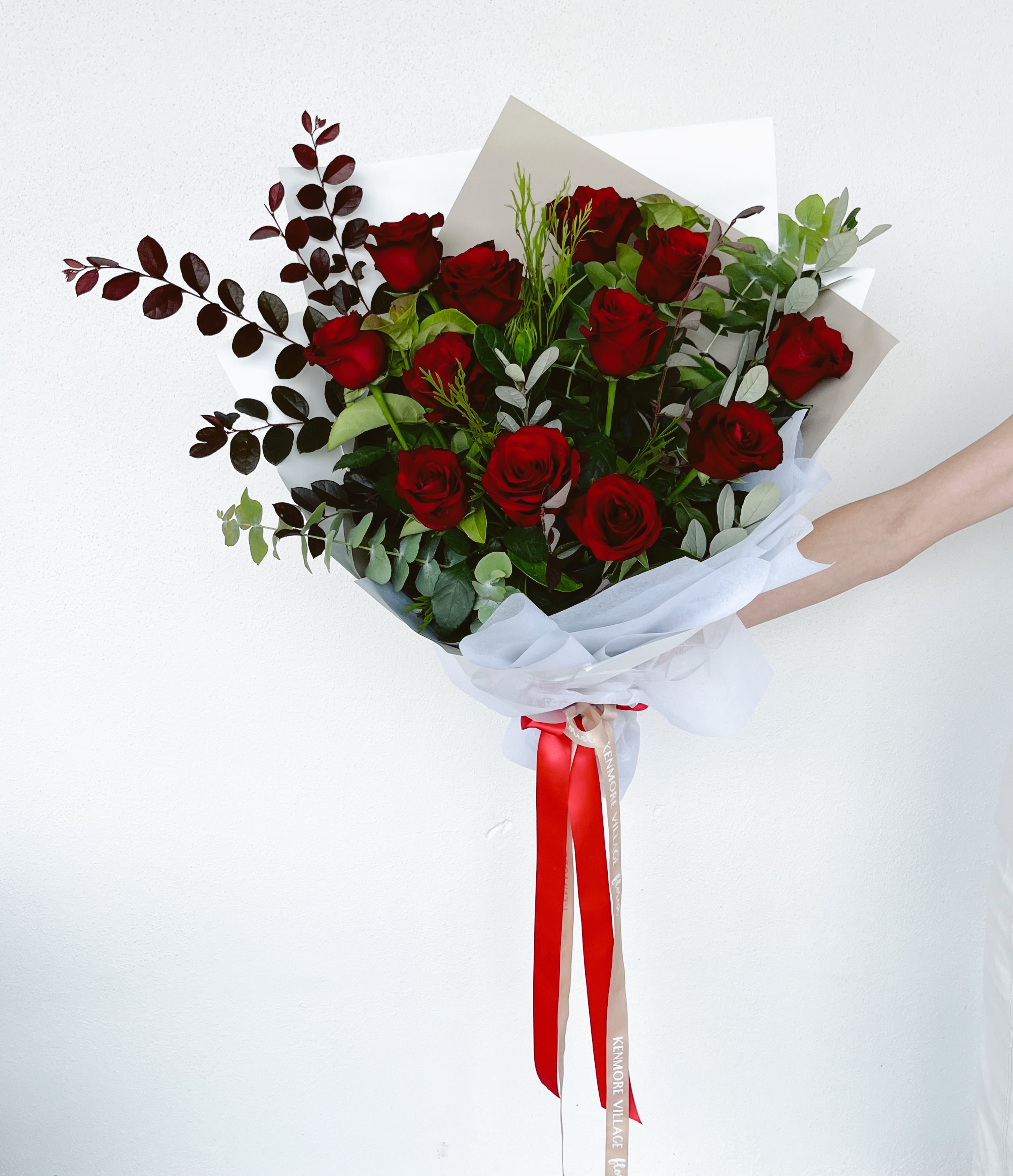 A dozen red roses with green leaves and decorative red ribbon, wrapped in a white paper wrap. Kenmore Village Florist. Brisbane Flower Delivery.