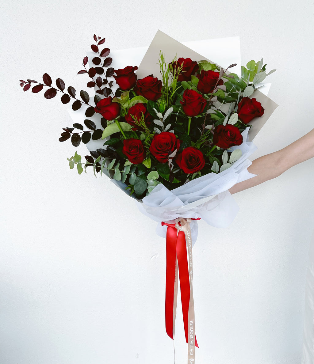 A dozen red roses with green leaves and decorative red ribbon, wrapped in a white paper wrap. Kenmore Village Florist. Brisbane Flower Delivery.