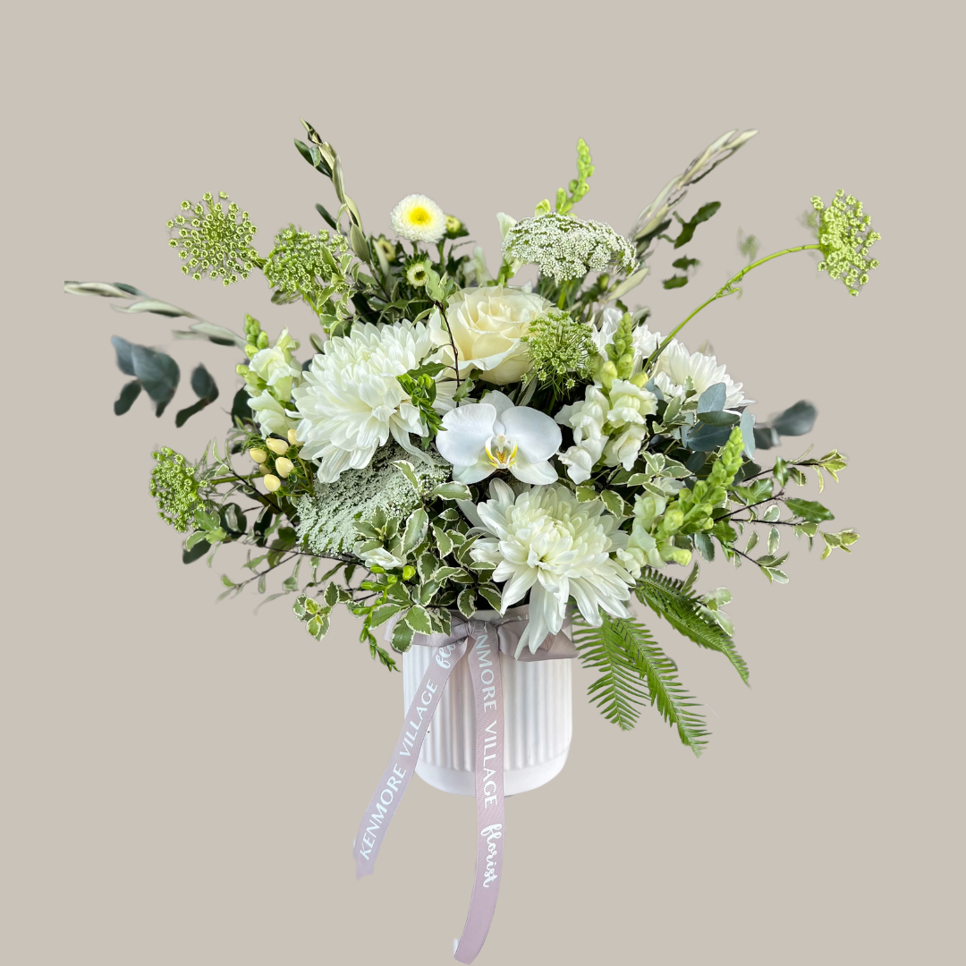A floral arrangement with a selection of soft white, cream, and green seasonal blooms, presented in a simple white ceramic pot, tied with a ribbon. Sympathy Flowers. Kenmore Village Florist. Brisbane Flower Delivery.