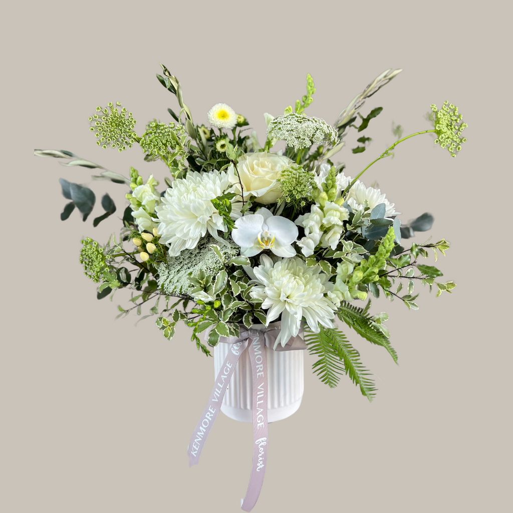 A floral arrangement with a selection of soft white, cream, and green seasonal blooms, presented in a simple white ceramic pot, tied with a ribbon. Sympathy Flowers. Kenmore Village Florist. Brisbane Flower Delivery.