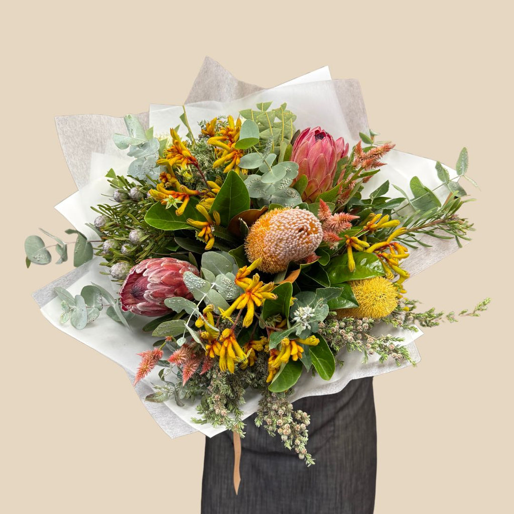 A bouquet of fresh mixed native flowers with green, yellow, and red colors, wrapped in white paper with ribbon. Brisbane Flower Delivery. Kenmore Village Florist. Same day delivery available.