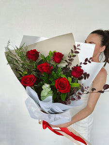 A half dozen red roses with green leaves and decorative red ribbon, wrapped in a white paper wrap.